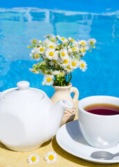 A cup of tea, a teapot and a vase with a bouquet of daisies on the background of the pool.