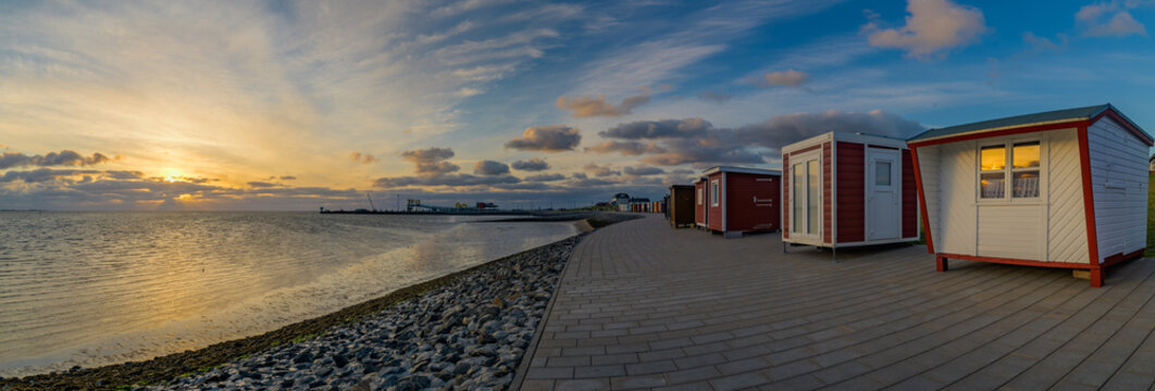 Panorama view of colorful beach houses and bathhouses on the dike of Dagebüll and its harbour on the background. Sunset reflection in the window
of beach house on the dike at the North Sea.

