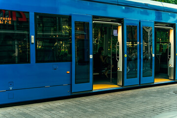 Blue tram with open doors for passengers at a stop on a summer evening, reflections in the windows...