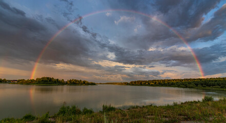 Picturesque rural landscape. Rainbow against a dramatic sky. Evening by the river.