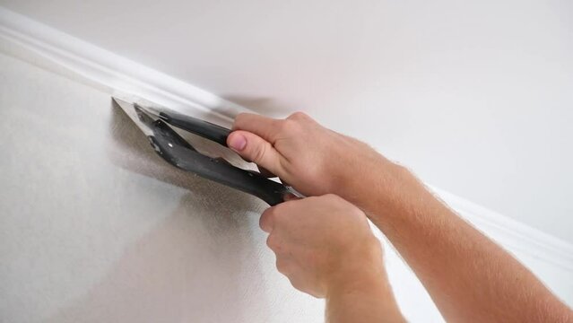 A man cuts wallpaper near ceiling moldings using box cutter and putty blade. Wallpapering. Repair.