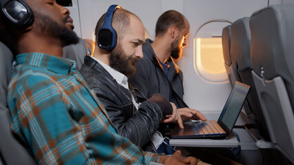 Businessman passenger using laptop computer on flight, travelling on work trip with tourists. Flying in economy class and working on device with online internet, sitting on airplane.