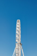 Side view of a tall white metal Ferris Wheel in front of a clear blue sky. Summer vacation advertisement or presentation background. 