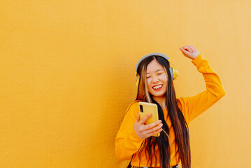 happy asian young woman in yellow sweater listening to music with a headphone and holding her...