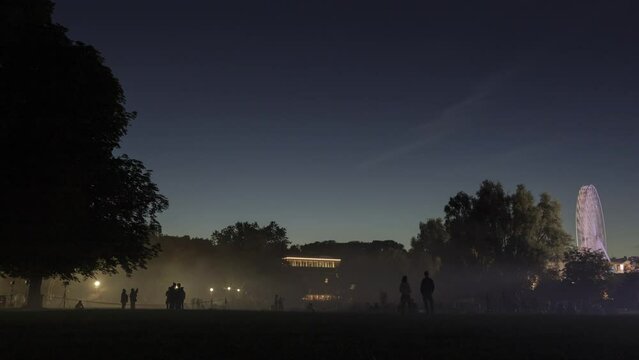 Time lapse shot of a laser show next to a fair with ferris wheel in background at night with the silhouettes of some people