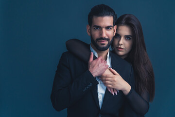 Hispanic man with beard wearing suit hugged by girlfriend from the back putting his hand on her arms looking seriously into camera. Dark background studio shot. High quality photo