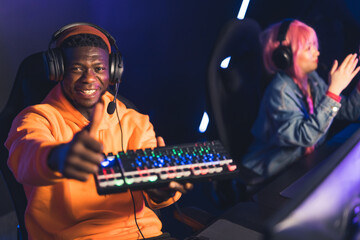 Young black man wearing orange hoodie celebrating win in online game tournament showing glowing keyboard up to the kamera and giving thumbs up. . High quality photo