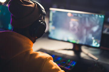 Teenager in orange clothes wearing headphones sitting in front of wide screen playing online game....