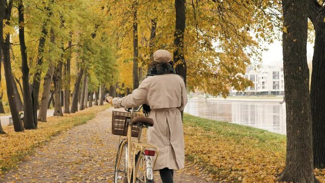 Young woman in trench coat and scarf walks holding bike handlebar against yellow trees on autumn park alley low angle shot
