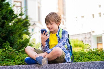 happy handsome boy with backpack sits in the park after school, calls parents or friends, communicates by video call, using mobile outside