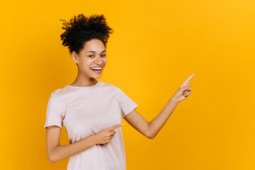 Cheerful positive lovely brunette millennial african american young woman, points with fingers to empty space, looks at camera, smiling friendly, stands on isolated orange background in casual t-shirt