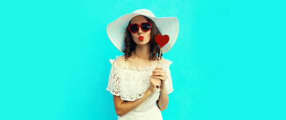 Portrait of beautiful young woman blowing her lips with red lipstick with sweet heart shaped lollipop wearing white summer straw hat, sunglasses on blue background