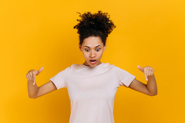 Stunned shocked african american curly haired young woman, surprised looks down and points fingers down, at empty copy space, stands on isolated orange color background in a basic t-shirt