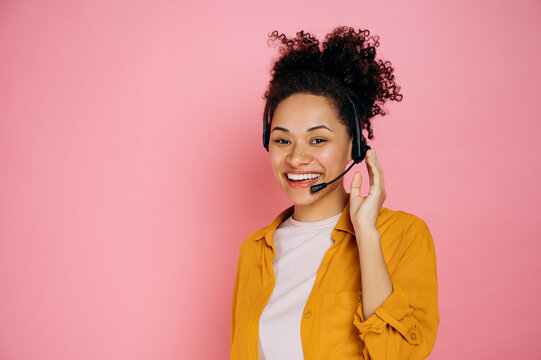 Happy african american curly haired young woman with headset, help desk worker, call center operator, consultant, talking with client, stand on isolated pink background, looks at camera,smile friendly