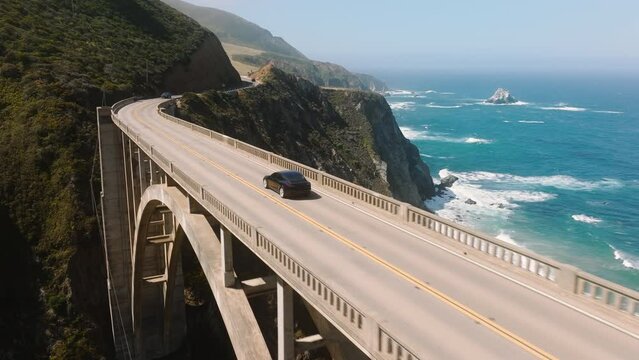 Electric car driving by scenic bridge on cinematic ocean background. Electric sedan drives by mountain coast road, scenic route. Alternative energy for cars. Eco and environmentally friendly transport
