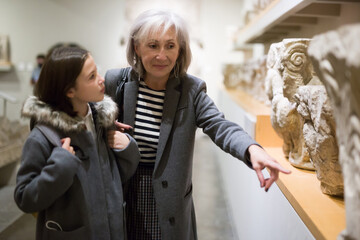Inquisitive preteen girl visiting museum of ancient sculpture with her grandmother, exploring antique showpieces together