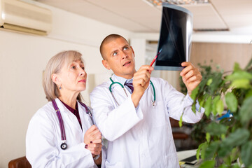 Obraz premium Experienced aged female doctor advising young male colleague about diagnosis of patient while checking x-ray at clinic office