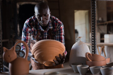 Hardworking African guy engaged in pottery, looking at clay dishware he created