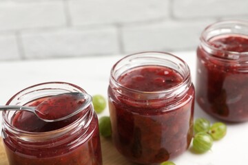 Jars of delicious gooseberry jam and fresh berries on white table, closeup