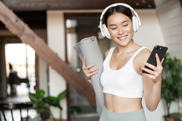 Smiling asian girl with headphones, looking at mobile phone and holding rubber mat for workout,...