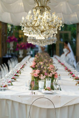 Wedding banquet table with pink rose flowers decoration, crystal chandelier in summer garden, selective soft focus, bride in white dress on background. Table set for event, party in outdoor restaurant