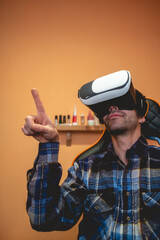 Handsome and happy young latino man with plaid shirt playing with virtual reality helmet (VR glasses) on a gamer chair in orange bedroom on home