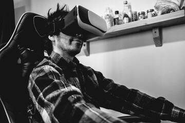 Handsome and happy young latino man with plaid shirt playing with virtual reality helmet (VR glasses) on a gamer chair in orange bedroom on home (in black and white)