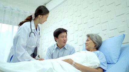 Asian man sitting beside his elder mother on the bed in hospital ward and doctor is coming to tell him about the medical analyze result.
