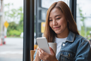 Fototapeta premium Portrait image of a young woman holding and using mobile phone while drinking coffee in cafe