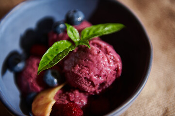 Refreshing raw vegan purple berry sorbet, on navy ceramic bowl, with lemon basil leaves, blueberries and juicy peaches.
