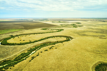 a winding muddy river with overgrown green banks in a sun-scorched steppe rural landscape