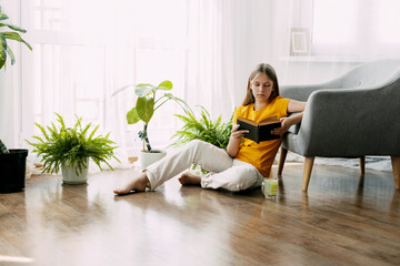 An attractive girl is reading a book, sitting on the floor of the house, having a good time. Rest,...
