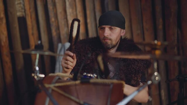 Bearded man with fur plays drums sitting near rough logs