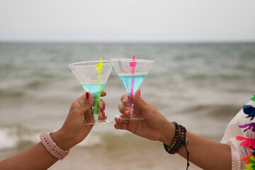 Detail of two hands raising glasses in the air. They are toasting for the good things that happen. The photo is taken from below and you can see the sky and the sea in the background.