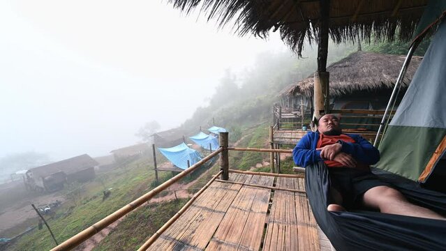 Asian fat traveler Sleeping on hammock on the wooden balcony with beautiful landscape and mist in the morning of Sapan Village nan Thailand.Sapan is Small and tranquil Village in the mountain.
