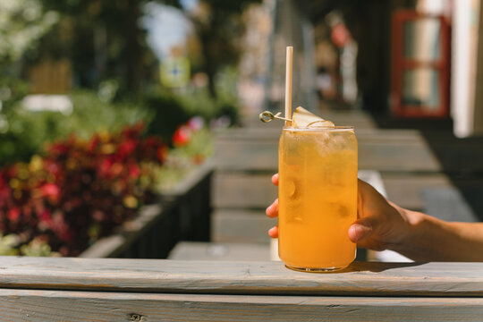 Close up of woman holding orange lemonade in glass outdoors. Summer refreshing drinks. Copy space. Blurred city on background.