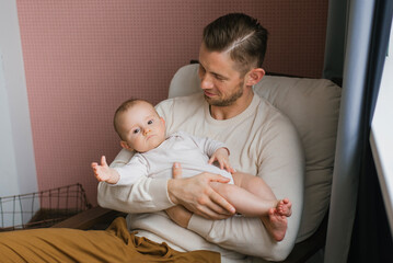 Portrait of a caring Caucasian dad holding a cute baby son in his arms, sitting in a comfortable chair at home