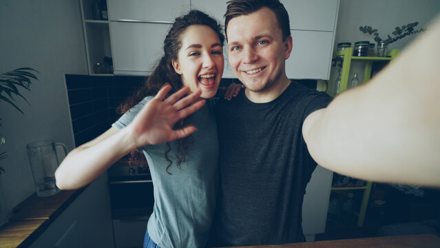 Point of view shot of young happy couple having online video call with smartphone camera while standing in the kitchen at home