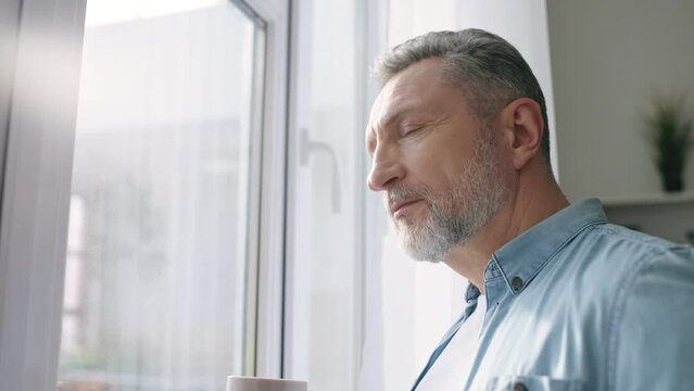 Thoughtful middle-aged man drinking morning coffee near window, midlife crisis