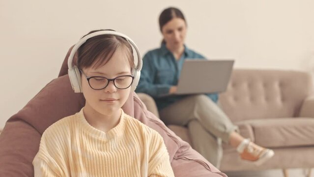 Smiling little girl with down syndrome listening to music in headphones at home while er mother browsing on laptop sitting on sofa in background