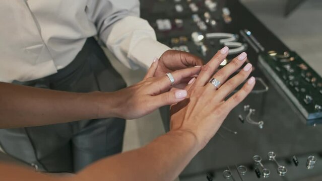 Top-down view of cropped woman standing by display in modern jewelry store, looking at rings on her hands, female consultant helping