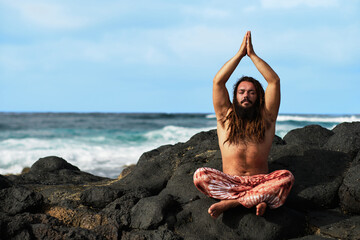 Rasta boy with beard doing yoga posture sitting with his hands together on his head. by the sea.