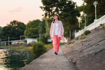 A young stylish caucasian woman in summer pink clothes and straw hat walking down at river...