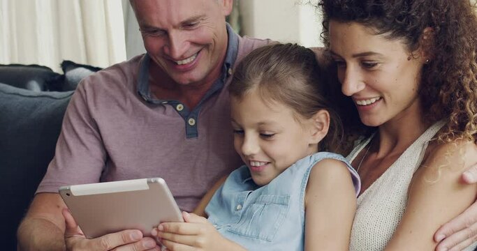 Happy and diverse foster family browsing on a tablet while relaxing in the living room at home. Married husband and wife with their adopted daughter smiling and surfing the internet over the weekend