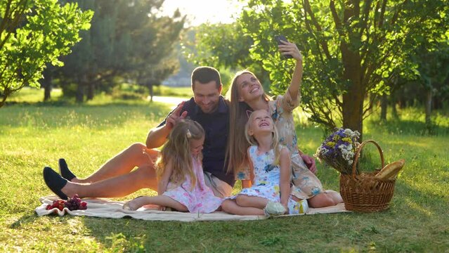 Happy family having picnic sitting on blanket in park on sunny day and taking selfie photos on smartphone. Young cheerful parents taking pictures with kids on mobile phone outdoor