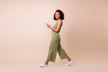 Photo of pretty cute lovely girl curly hairdo wear khaki overall white sneakers texting in smartphone isolated on beige color background