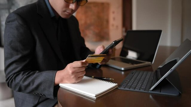 business Asian man hand using smart phone, tablet payments and holding credit card online shopping, omni channel, digital tablet docking keyboard computer at office in sun light.