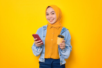 Smiling young Asian woman in jeans jacket holding mobile phone and coffee cup isolated over yellow background