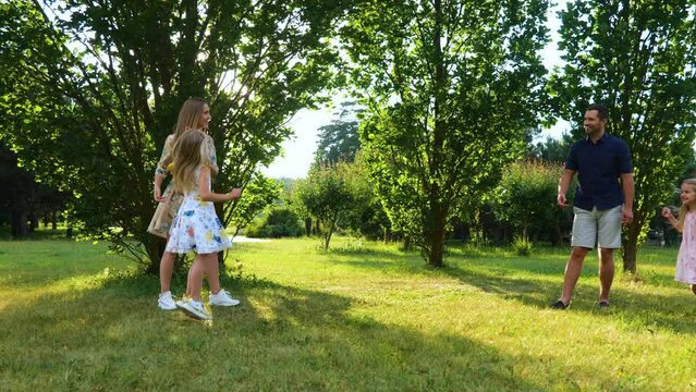 Caucasian Family Throwing Frisbee Disk In Park Outside. Happy Girls Jumping Cheering For Parents While Mom And Dad Play With Frisbee Plate. Children And Parents. Outdoor Activity Concept