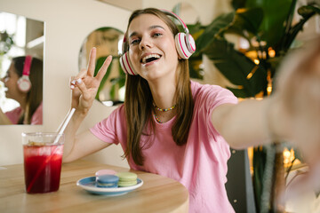 Close portrait of a young teen girl in headphones makes a selfie on the phone. cute Brunette sits...